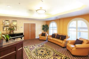 A cozy lounge with tan sofas and dark cushions, a grand piano, and potted plants on a purple-patterned carpet.