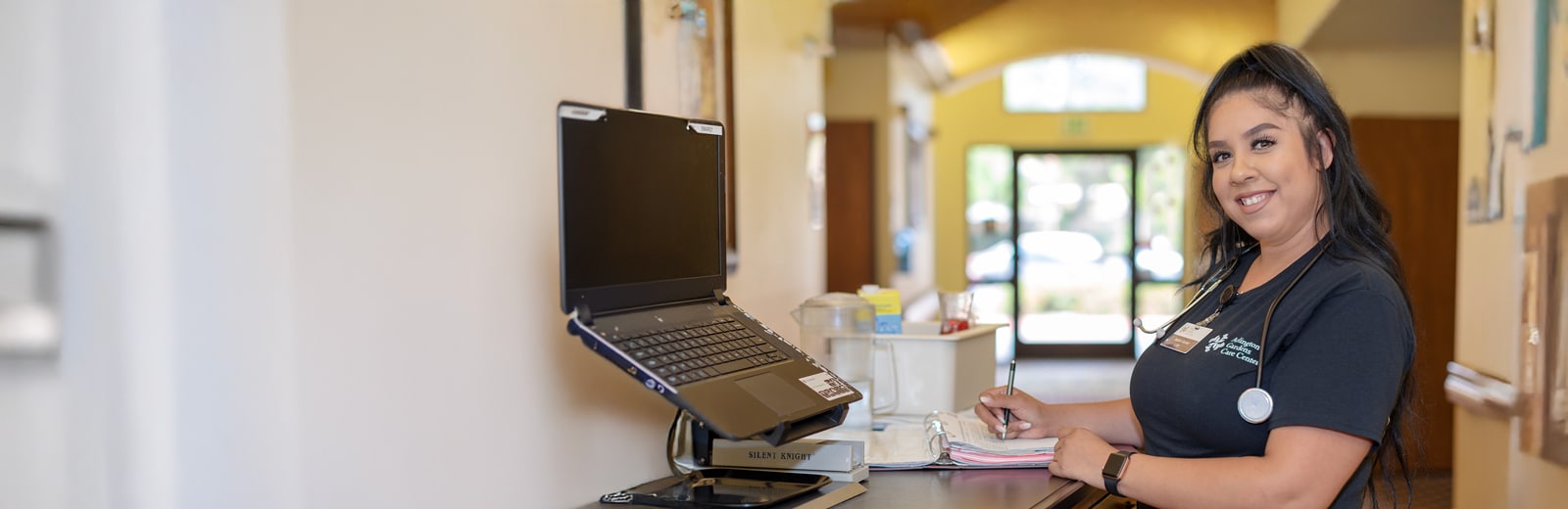 Smiling nurse with a stethoscope writing in a folder at a reception desk. The background shows a bright hallway with open doors
