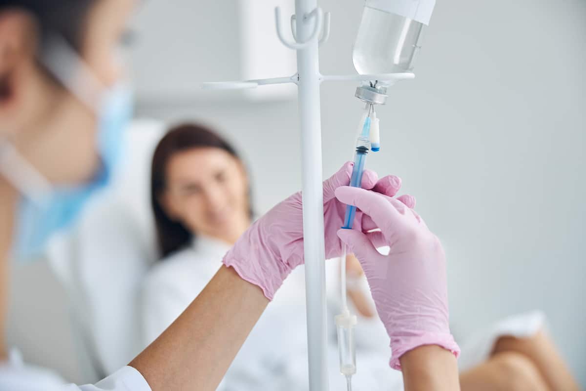 A healthcare professional in pink gloves adjusts an IV drip, with a blurred patient sitting in the background