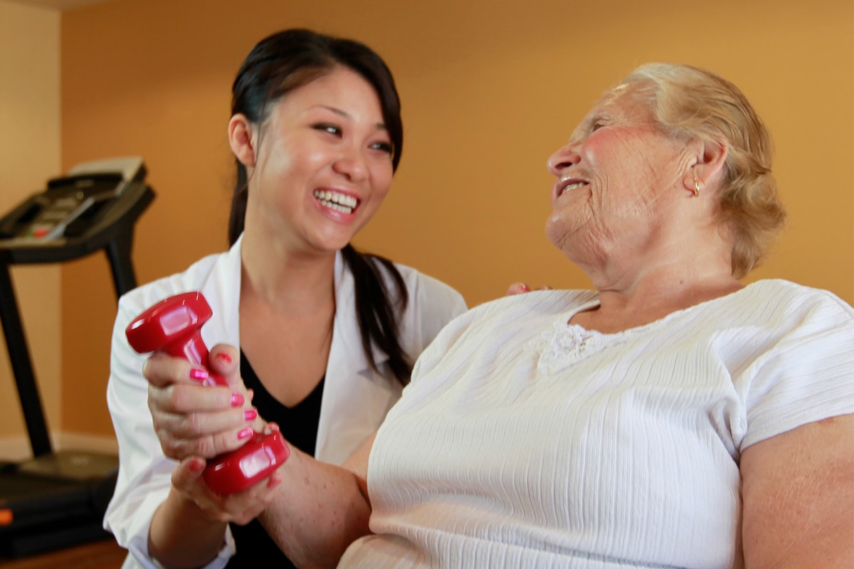 A healthcare worker assists an elderly woman with a red dumbbell, both smiling warmly.