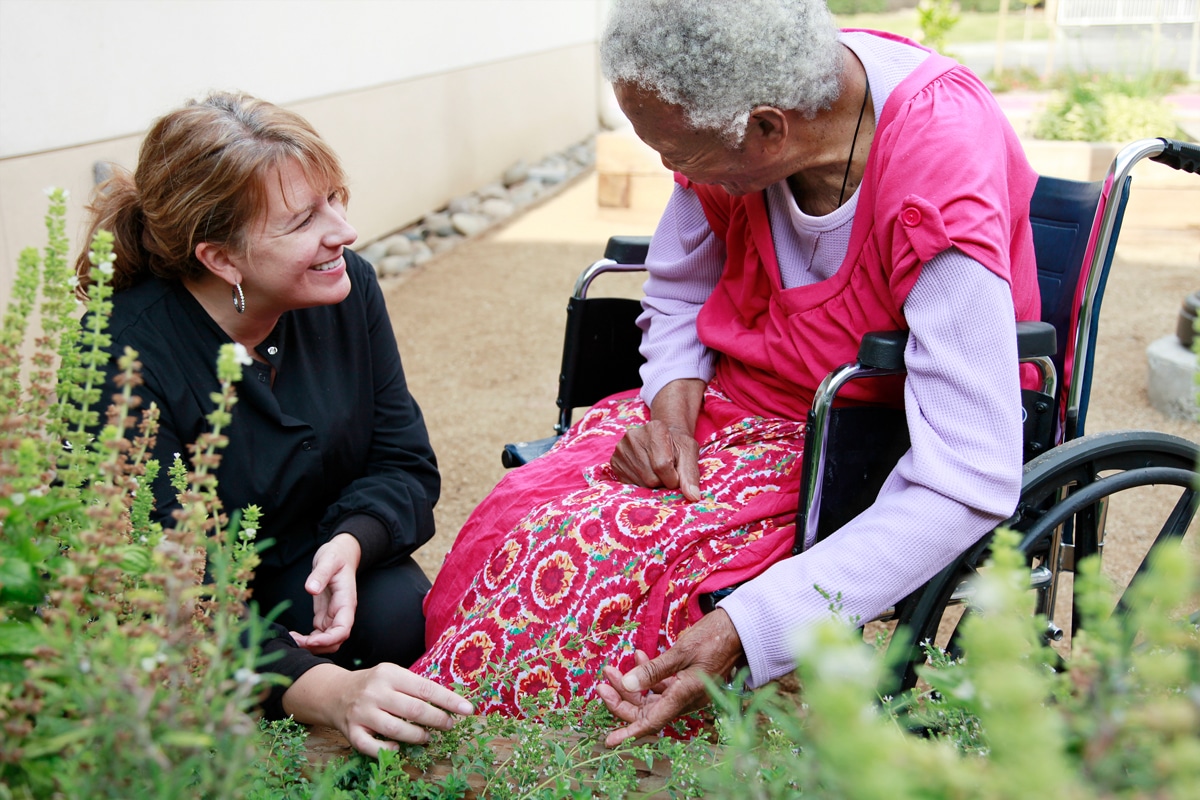 A nurse is sitting beside an elderly woman in a wheelchair in a garden. They smile warmly at each other, surrounded by lush plants.