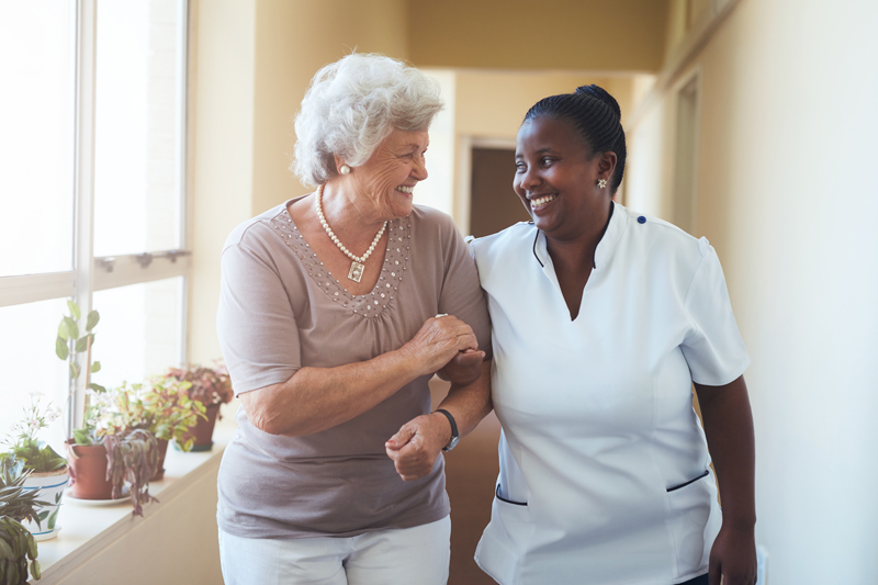 Elderly woman and caregiver walk arm-in-arm down a sunlit hallway. Both are smiling, conveying warmth and companionship.