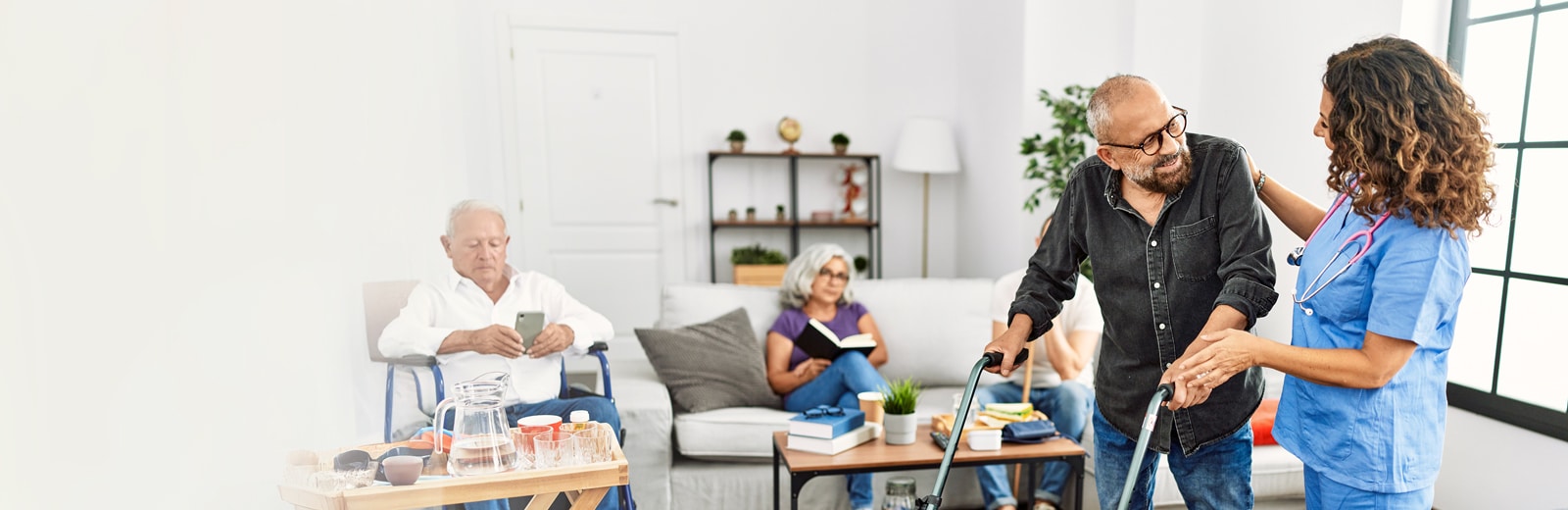 A nurse assists an elderly man with a walker in a cozy room. Other seniors are seated, reading or using a phone.