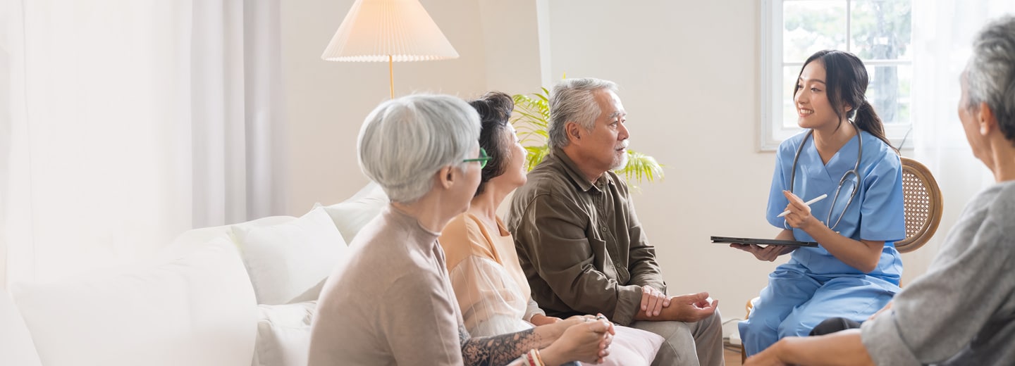 A nurse in blue scrubs smiles and talks with a group of elderly people in a bright living room.